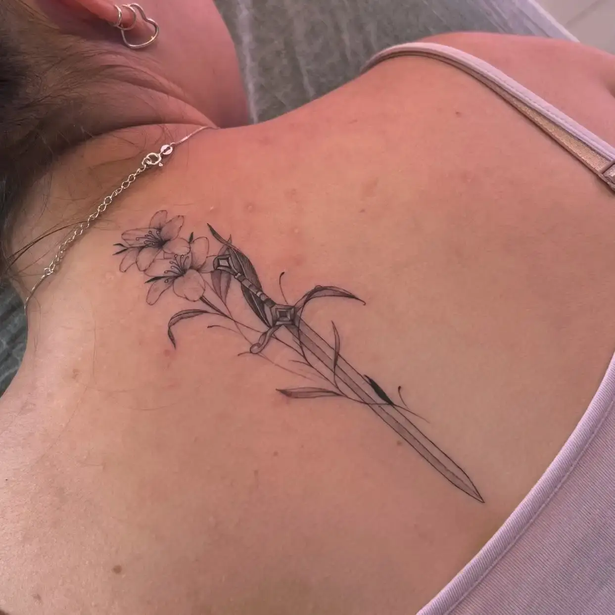 Fine-line lily and dagger upper back tattoo by Paulo Rodrigues at XOXO Dublin